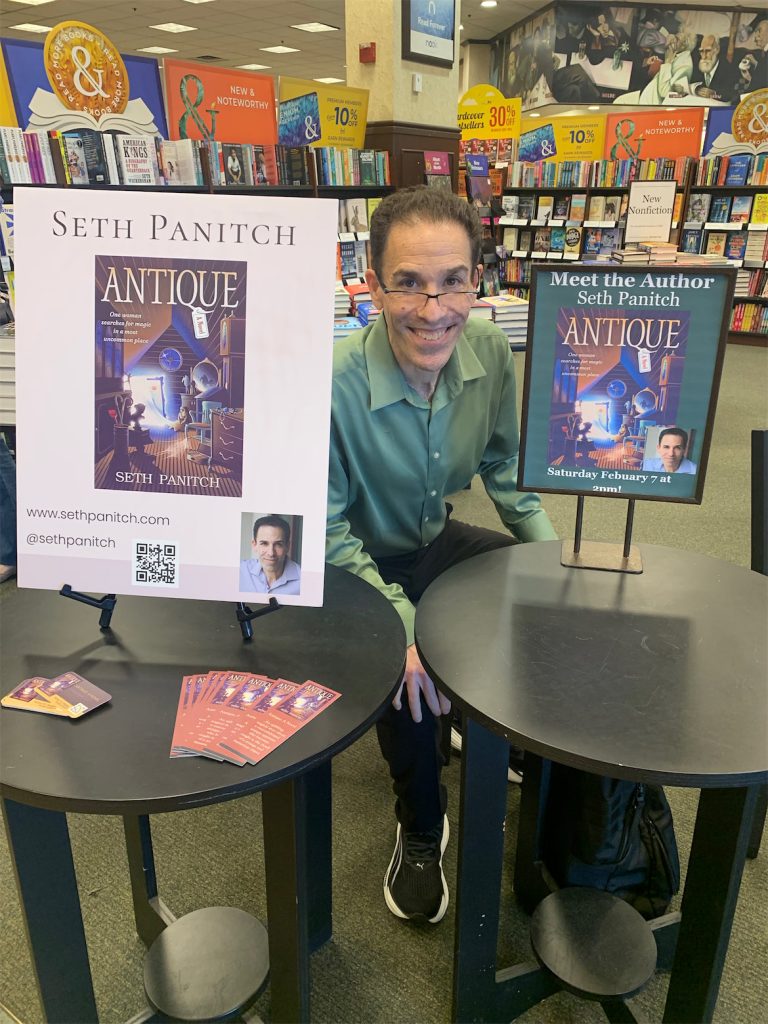 Seth Panitch sitting at table smiling at camera next to two ad displays for his book antique.